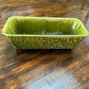 May include: A rectangular, olive green ceramic dish with a textured, bubble-like pattern on the sides. The dish has a glossy finish and is likely used for serving or display. The dish is on a wooden surface.
