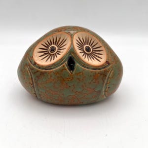 May include: A rounded ceramic owl figurine featuring a mottled green and brown glaze. The owl has two large, light-colored circular eye patches with dark, radiating lines and small black pupils, along with a small, dark, triangular beak.