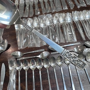 May include: A stainless steel tablespoon held in the foreground, featuring a decorative embossed pattern on the handle. Numerous matching spoons, some individually wrapped in clear plastic, are arranged on a dark wooden surface below.