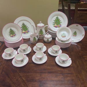 May include: A complete Christmas-themed dinnerware set. The white porcelain pieces feature a green Christmas tree design with red trim. The set includes plates, bowls, cups, saucers, a teapot, creamer, sugar bowl, and serving dishes.