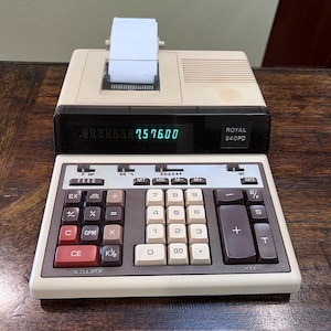May include: A vintage Royal 340PD calculator with a built-in printer. The calculator has a beige and black color scheme with a green LED display showing the number 7576.00. The keyboard features various function keys.