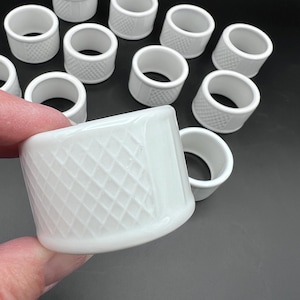 May include: A collection of white ceramic thimbles, some with a diamond pattern. The thimbles are cylindrical with a rounded top and a hole for the finger. The thimbles are on a dark surface.
