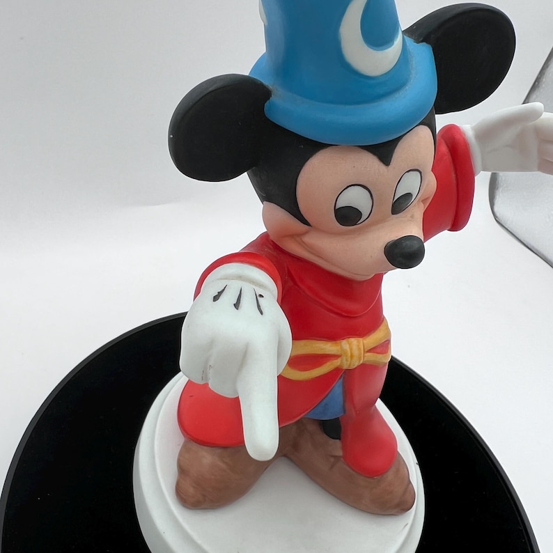 May include: A Mickey Mouse figurine dressed as a sorcerer, wearing a blue hat with a crescent moon, a red robe, and white gloves. The figurine is standing on a white base and pointing with one hand.