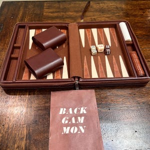 May include: A brown leather backgammon set featuring a folding case with a felt interior, light and dark brown triangular points, two dice cups, white and brown checkers, two standard dice, and a doubling cube with the number 64 printed on it.