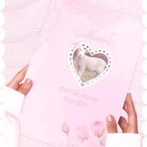May include: A light pink medication tracker notebook featuring a white heart-shaped frame with a small lamb inside, a pink bow, and the text "Maries's medication tracker" printed in a script font, accented with floral rose illustrations at the bottom.