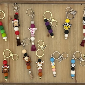 May include: A collection of 12 colorful beaded keychains with various charms, including a Christmas tree, a cow, a smiley face, and a daisy. Each keychain features a gold metal ring and a tassel.