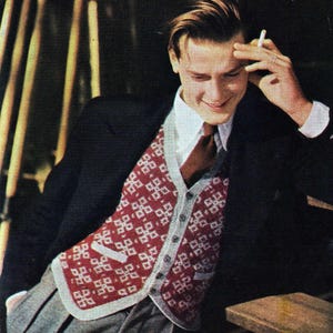 May include: A man in a black blazer, white shirt, and a red and white patterned vest. He wears a brown tie and grey trousers. He is holding a cigarette and smiling.