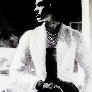May include: A woman wearing a white fur jacket with a black dress and a pearl necklace. The woman is looking to the left of the image.