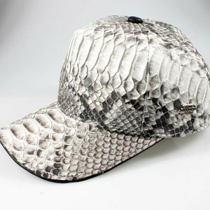 以下が含まれることがあります： 白とグレーのヘビ革製のベースボールキャップ。黒いつばと、後ろに銀色の金属製のロゴが付いている。