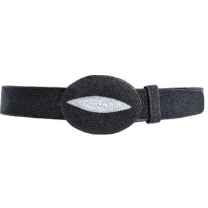 Puede incluir: Un cinturón de cuero negro con una hebilla ovalada blanca. La hebilla tiene un diseño único que se asemeja a un ojo.
