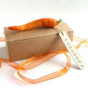 May include: A small brown cardboard box with a felt carrot on top. The box has a brown paper tag attached to it that says "CARROT". The box is sitting on a white surface with an orange ribbon.