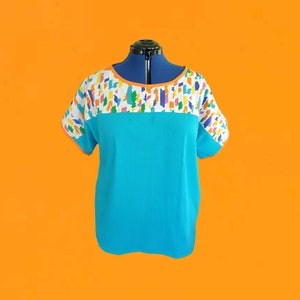 May include: A turquoise short-sleeved top with a color block design. The top features a white yoke with a colorful geometric pattern and an orange neckline. The fabric has a textured appearance.