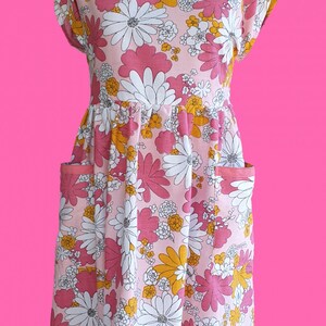 May include: A pink floral print dress with white, yellow, and pink flowers. The dress has a round neckline, short sleeves, and two front pockets. The dress is displayed on a red dress form against a bright pink background.