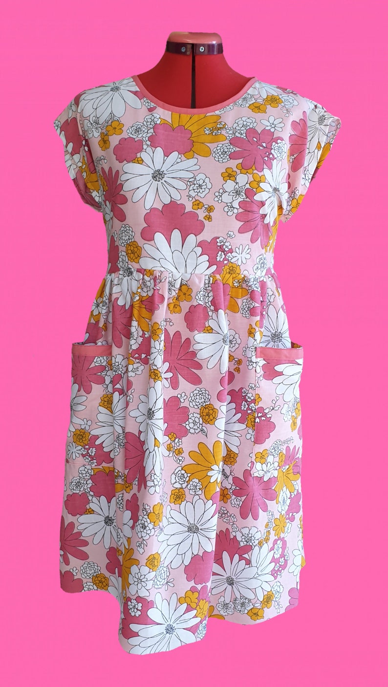 May include: A pink floral print dress with white, yellow, and pink flowers. The dress has a round neckline, short sleeves, and two front pockets. The dress is displayed on a red dress form against a bright pink background.