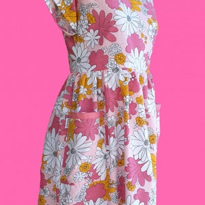May include: A pink floral print dress with white, yellow, and pink flowers. The dress has a round neckline, short sleeves, and two pockets. The dress is displayed on a red dress form against a bright pink background.