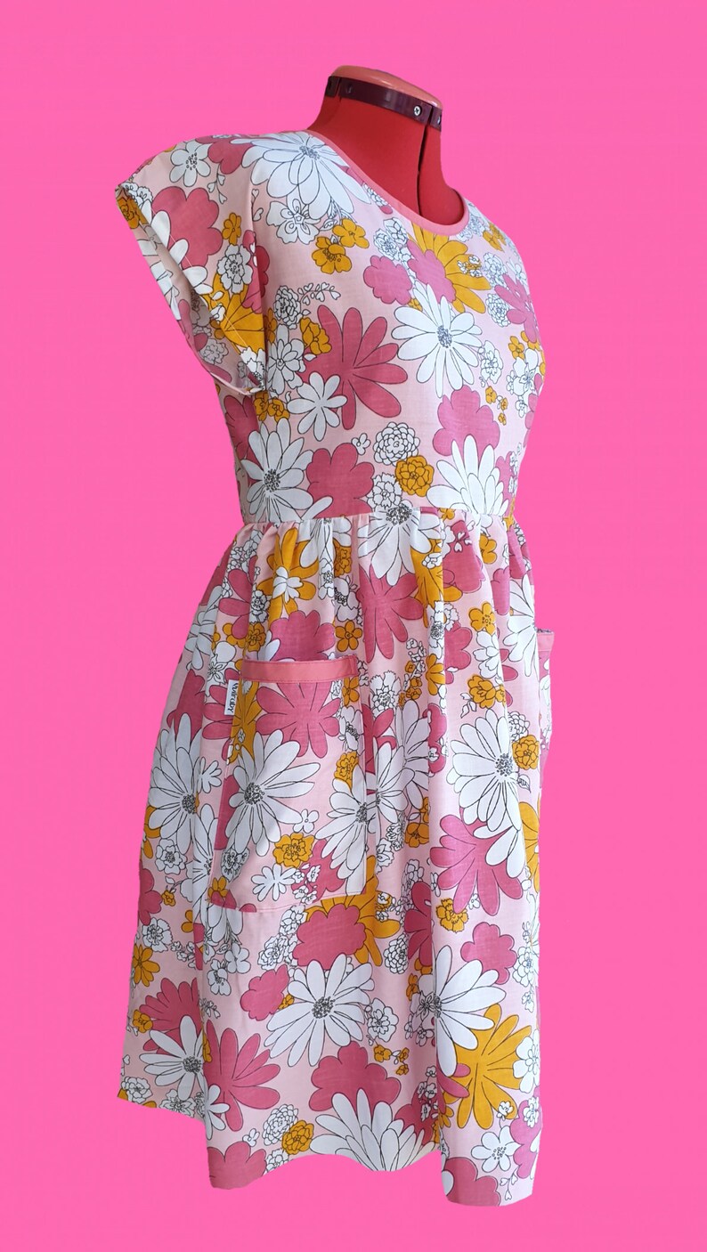 May include: A pink floral print dress with white, yellow, and pink flowers. The dress has a round neckline, short sleeves, and two pockets. The dress is displayed on a red dress form against a bright pink background.