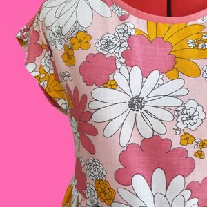 May include: A pink dress with a large floral pattern featuring white, pink, and yellow flowers. The dress has short sleeves and a round neckline with a pink trim. The fabric appears to be cotton or a similar material.