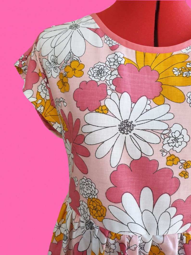 May include: A pink dress with a large floral pattern featuring white, pink, and yellow flowers. The dress has short sleeves and a round neckline with a pink trim. The fabric appears to be cotton or a similar material.