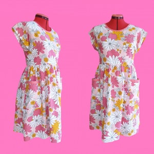 May include: A light pink floral print dress with white, yellow, and pink flowers. The dress has short sleeves, a round neckline, and two front pockets. The dress is displayed on a red dress form against a pink background.