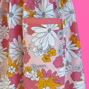 May include: A close-up of a pink floral apron with a pocket. The apron features a large-scale floral pattern in shades of pink, white, and yellow. The pocket is a solid coral color. The apron is made of a lightweight fabric.