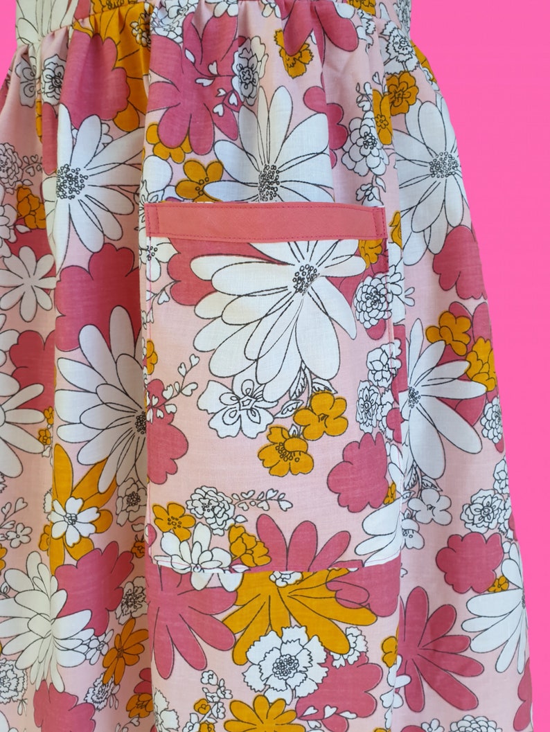 May include: A close-up of a pink floral apron with a pocket. The apron features a large-scale floral pattern in shades of pink, white, and yellow. The pocket is a solid coral color. The apron is made of a lightweight fabric.