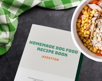 May include: A white book with the title "Homemade Dog Food Recipe Book" in green and orange text. The book is on a dark gray surface with a green and white checkered cloth in the background. A white bowl with a dog food recipe is in the top right corner of the image.