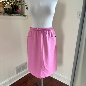 May include: A pink knee-length skirt with two front pockets and a drawstring waist. The skirt is displayed on a white mannequin. The skirt is made of a smooth, solid-coloured fabric.
