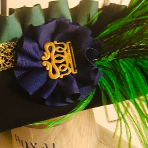 Pu&ograve; includere: Un cappello verde scuro con un fiore di velluto blu e un accento di piuma verde. Il fiore ha un ornamento in metallo dorato con un design decorativo.
