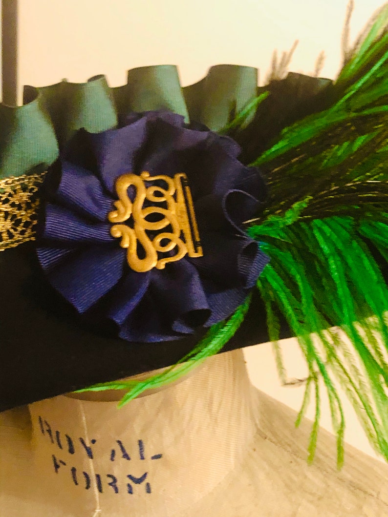 Pu&ograve; includere: Un cappello verde scuro con un fiore di velluto blu e un accento di piuma verde. Il fiore ha un ornamento in metallo dorato con un design decorativo.