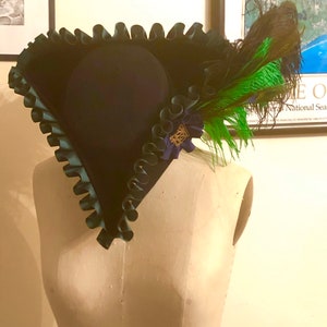 Pu&ograve; includere: Un cappello blu scuro con un bordo verde a balze e un accento di piuma verde e marrone.