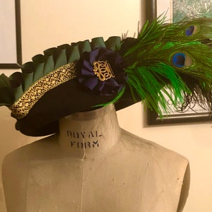 Pu&ograve; includere: Un cappello nero con un bordo verde e oro e un fiore blu con un accento dorato. Il cappello &egrave; adornato con una grande piuma di pavone verde e nera.