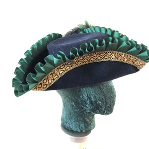 Pu&ograve; includere: Un cappello tricorno in feltro blu con un bordo arricciato verde e una treccia dorata. Il cappello ha una piuma in cima.