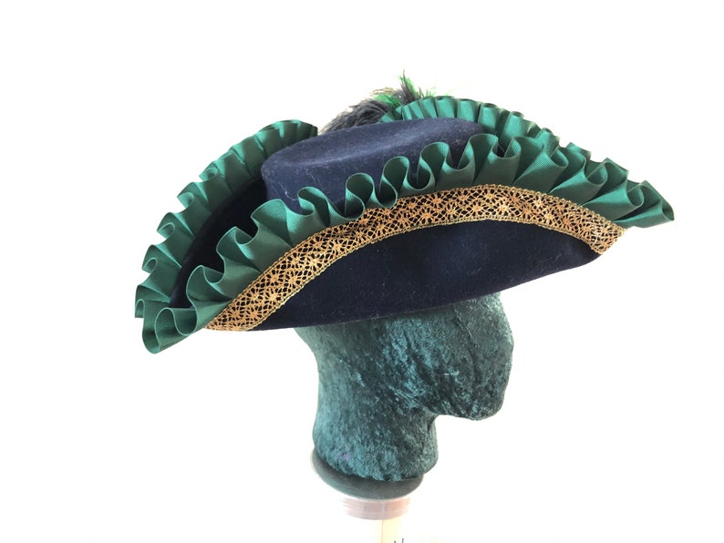 Pu&ograve; includere: Un cappello tricorno in feltro blu con un bordo arricciato verde e una treccia dorata. Il cappello ha una piuma in cima.