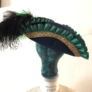 Pu&ograve; includere: Un cappello tricorno blu navy con un bordo verde e oro. Il cappello ha una piuma nera.