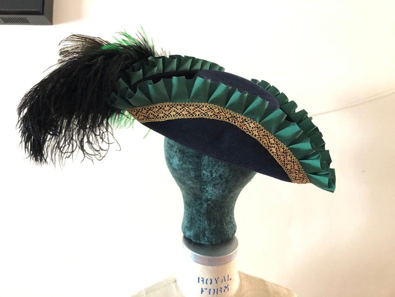 Pu&ograve; includere: Un cappello tricorno blu navy con un bordo verde e oro. Il cappello ha una piuma nera.