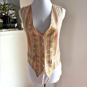May include: A vintage vest with a floral and striped pattern in shades of pink, gold, and green. The vest has a V-neck, button closure, and two small pockets. The vest is displayed on a mannequin.