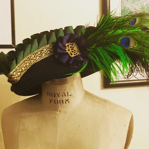 Pu&ograve; includere: Un cappello di feltro nero con un bordo verde a balze e una treccia dorata. Un fiore viola con un emblema dorato &egrave; attaccato al cappello. Due piume di pavone verdi sono attaccate alla parte posteriore del cappello.