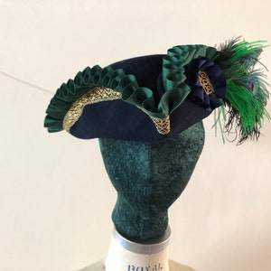 Pu&ograve; includere: Un cappello di feltro blu navy con un bordo verde e oro. Il cappello ha una piuma verde e un fiore blu sul lato.
