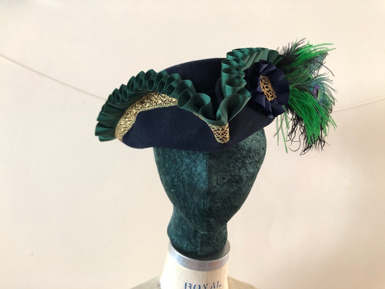 Pu&ograve; includere: Un cappello di feltro blu navy con un bordo verde e oro. Il cappello ha una piuma verde e un fiore blu sul lato.