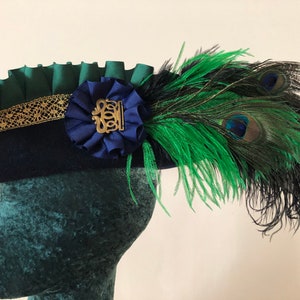 Pu&ograve; includere: Un cappello blu navy con un volant verde e un bordo dorato. Un nastro blu con una fibbia dorata &egrave; attaccato al cappello. Una piuma di pavone verde e nera &egrave; attaccata al lato del cappello.