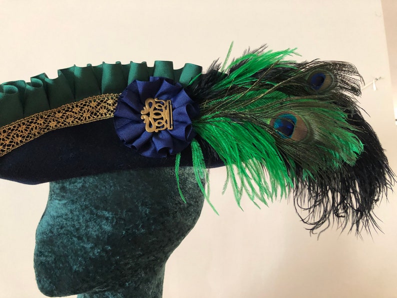 Pu&ograve; includere: Un cappello blu navy con un volant verde e un bordo dorato. Un nastro blu con una fibbia dorata &egrave; attaccato al cappello. Una piuma di pavone verde e nera &egrave; attaccata al lato del cappello.