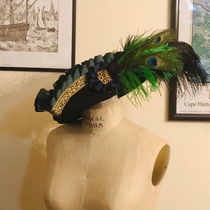 Pu&ograve; includere: Un cappello blu scuro con un bordo dorato e una piuma di pavone verde e blu. Il cappello &egrave; su un manichino bianco.