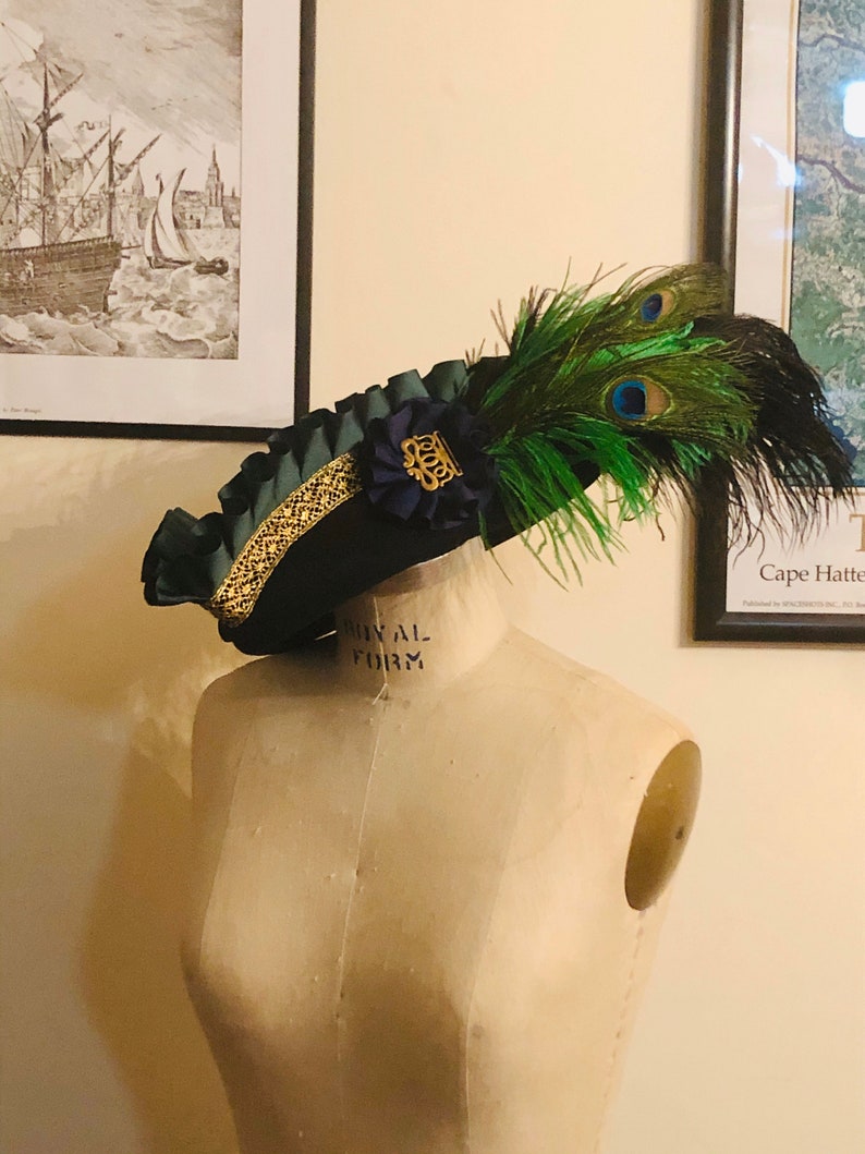 Pu&ograve; includere: Un cappello blu scuro con un bordo dorato e una piuma di pavone verde e blu. Il cappello &egrave; su un manichino bianco.