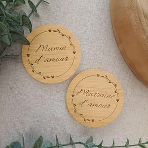 Puede incluir: Dos posavasos redondos de madera clara con texto grabado y un borde decorativo de corazones y hojas. Uno de los posavasos dice "Mamie d'amour" y el otro "Marraine d'amour".