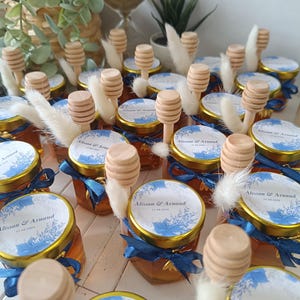 Peut inclure: Une collection de petits pots en verre remplis de miel, chacun avec une étiquette bleue et blanche qui indique "Alisson & Arnaud" et un petit bâtonnet en bois pour le miel. Les pots sont attachés avec un ruban bleu et décorés d'un petit charme d'abeille dorée.