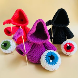 Peut inclure: Trois figurines à capuche crochetées avec des ornements en forme d'œil. Les figurines sont violettes, rouges et noires. Chaque figurine a un petit drapeau attaché à un bâton.