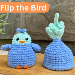 May include: Two handmade crochet figures: a blue and light blue bird and a blue figure with a light green hand giving the middle finger. The text "Flip the Bird" is in an orange rectangle above the figures. The figures are on a wooden cutting board.