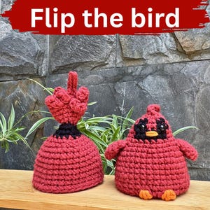 May include: Two handmade red crochet birds, one with a raised middle finger and the other with a cute face. The text "Flip the bird" is written in white on a red banner above the birds. The birds are on a wooden shelf.