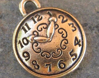 May include: Gold-toned metal charm shaped like a clock face with Roman numeral markings and a stylized hand pointing to the number 12. The charm has a loop at the top for attaching to a necklace or bracelet.