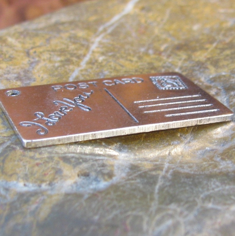 May include: A silver metal charm shaped like a postcard with the words "Post Card" and "Love You" engraved on it.